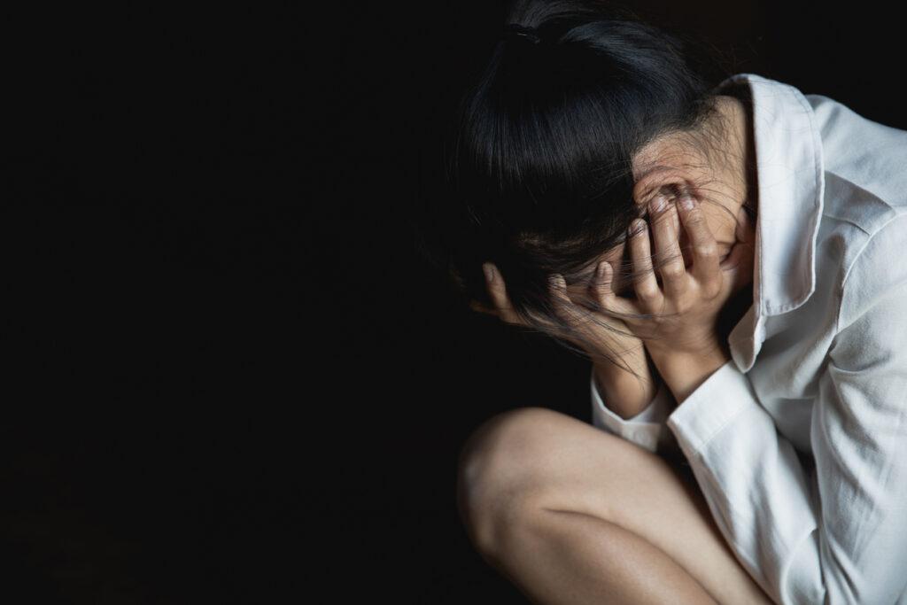 A depressed woman sitting alone in her room. / A woman in despair, covering her face in the dark.