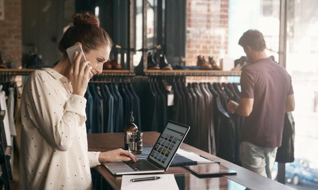 Woman using her phone and laptop in a clothing boutique.