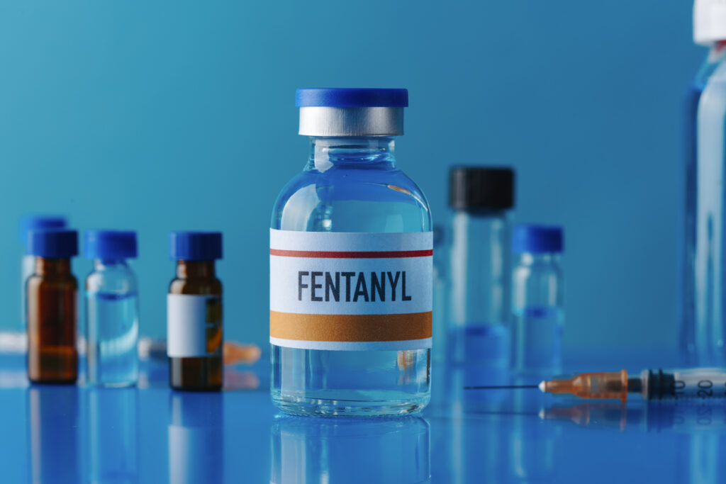 A simulated vial of fentanyl on a blue table next to a syringe.