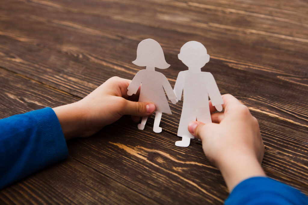 Child’s hands holding a  boy and a girl cardboard dolls on a wooden background.
